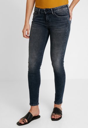 Kvinde iført falmede sorte skinny jeans og sorte slip-on sandaler, stående med den ene hånd hvilende på hoften.