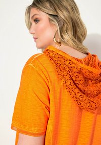 Oranger Spitzenkapuzenpullover mit kurzen Ärmeln, der eine strukturierte Spitzenkapuze und glattes Material aufweist und eine lebendige, einfarbige Optik ohne Muster bietet.