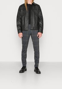 Zwarte faux leren jas met twee borstzakken, gecombineerd met grijze geruite broek en zwarte veterschoenen op een witte achtergrond.