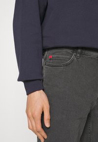 Mörkblå sweatshirt med ribbade muddknäppningar, tillsammans med grå jeans som har en röd flagga på fickan, vilket betonar textur och färgkontrast.