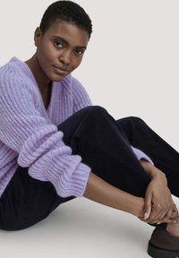 Hellblauer Strickpullover mit tiefem V-Ausschnitt und gerippten Bündchen, kombiniert mit dunklen Cordhosen. Schlichte braune Schuhe, sitzende Pose.