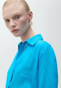 Camicia di lino blu brillante con colletto, caratterizzata da una superficie leggermente strutturata. Il modello indossa un orecchino a cerchio, lo sfondo è bianco semplice.
