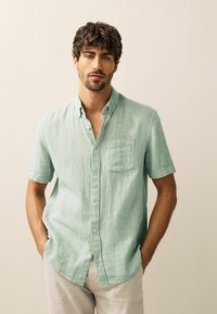 Short-sleeved, mint green button-up shirt made of textured fabric, featuring a chest pocket and a collar, worn with light-colored pants.