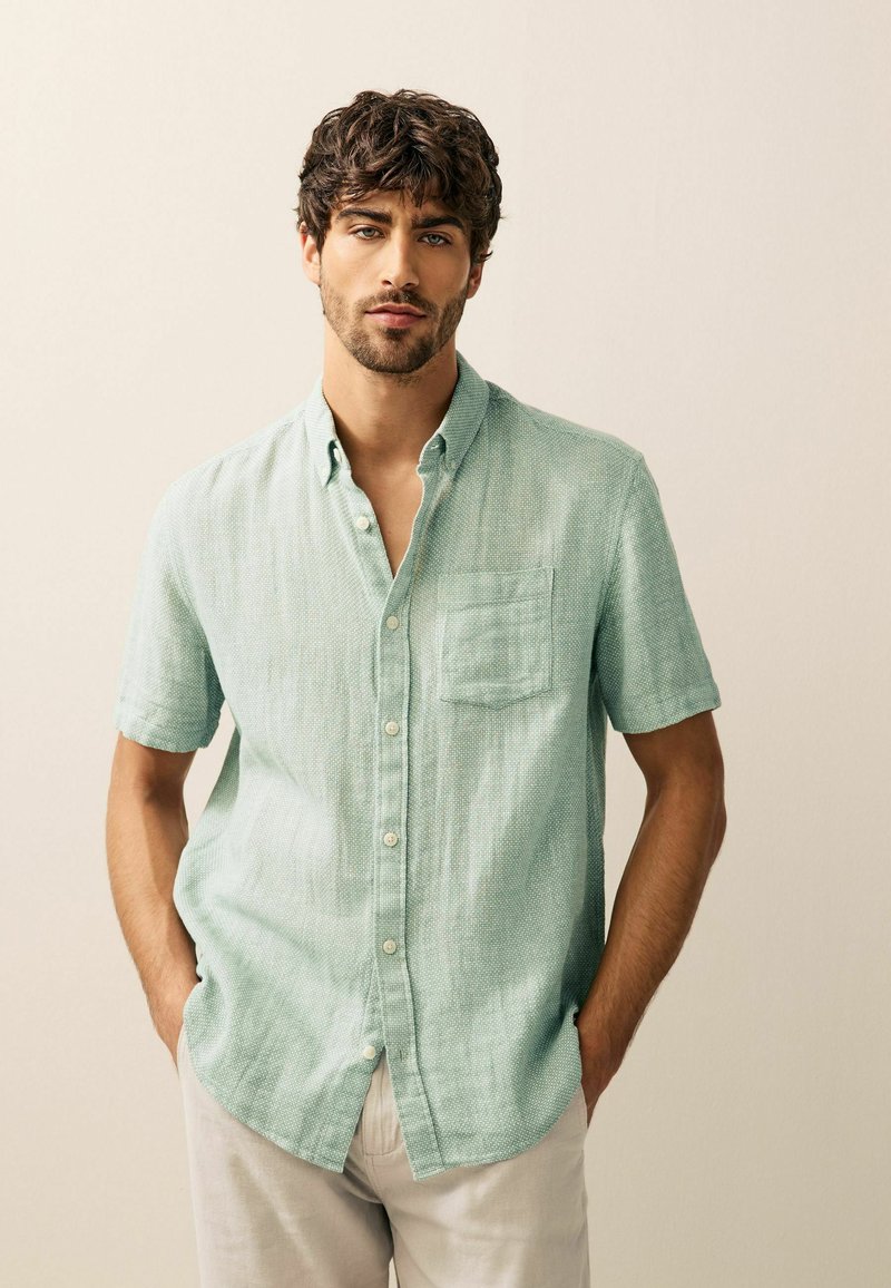 Short-sleeved, mint green button-up shirt made of textured fabric, featuring a chest pocket and a collar, worn with light-colored pants.