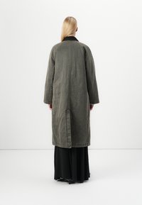 Cappotto lungo verde oliva con colletto nero e taglio dritto. Presenta una cucitura centrale posteriore e una vestibilità comoda. Indossato sopra un abito nero.