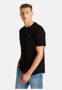 Schwarzes T-Shirt mit kurzen Ärmeln und rundem Ausschnitt, versehen mit einem kleinen Logopatch. Das Material sieht weich aus; getragen mit blauen Jeans.