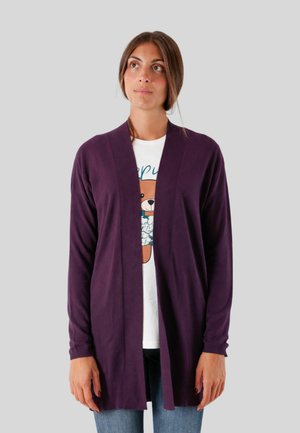 Donna con capelli castani che indossa un cardigan viola aperto davanti sopra una t-shirt bianca con grafica di un orsetto e jeans blu, in piedi davanti a uno sfondo grigio.