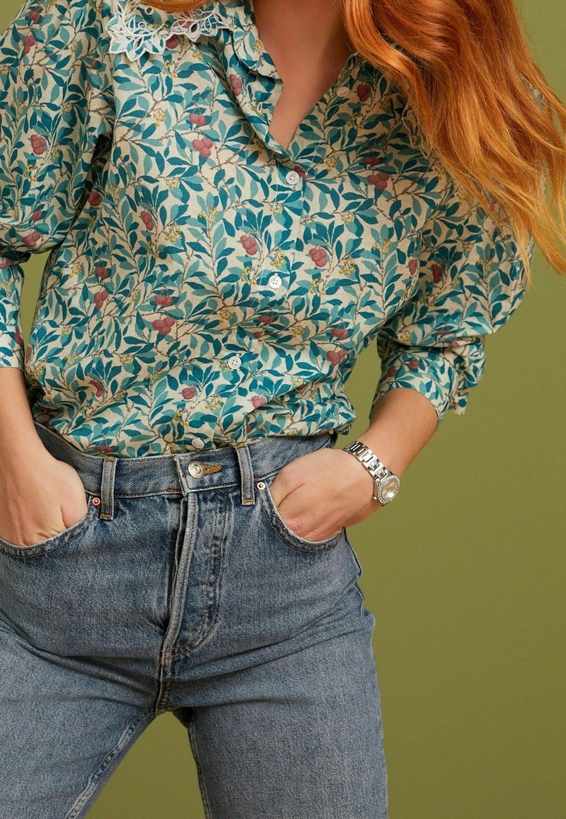 Chemise à motif floral bleu et crème avec un col en dentelle, associée à un jean en denim bleu taille haute. Montre en argent au poignet.