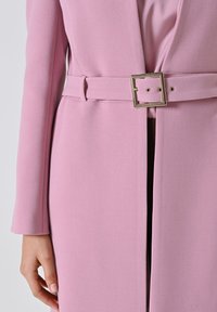 Cappotto su misura rosa chiaro con una texture liscia, caratterizzato da una vita con cintura e una fibbia metallica quadrata. Maniche lunghe e design pulito.