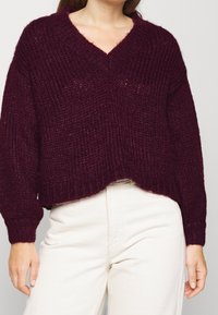 Burgundy stickad tröja med V-ringning, lös passform, ribbad struktur och långa ärmar, som matchas med ljusa byxor.