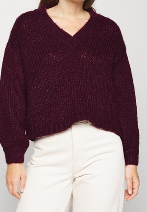 Burgunderfarbener Strickpullover mit V-Ausschnitt, lockerer Passform, gerippter Struktur und langen Ärmeln, kombiniert mit hellfarbenen Hosen.