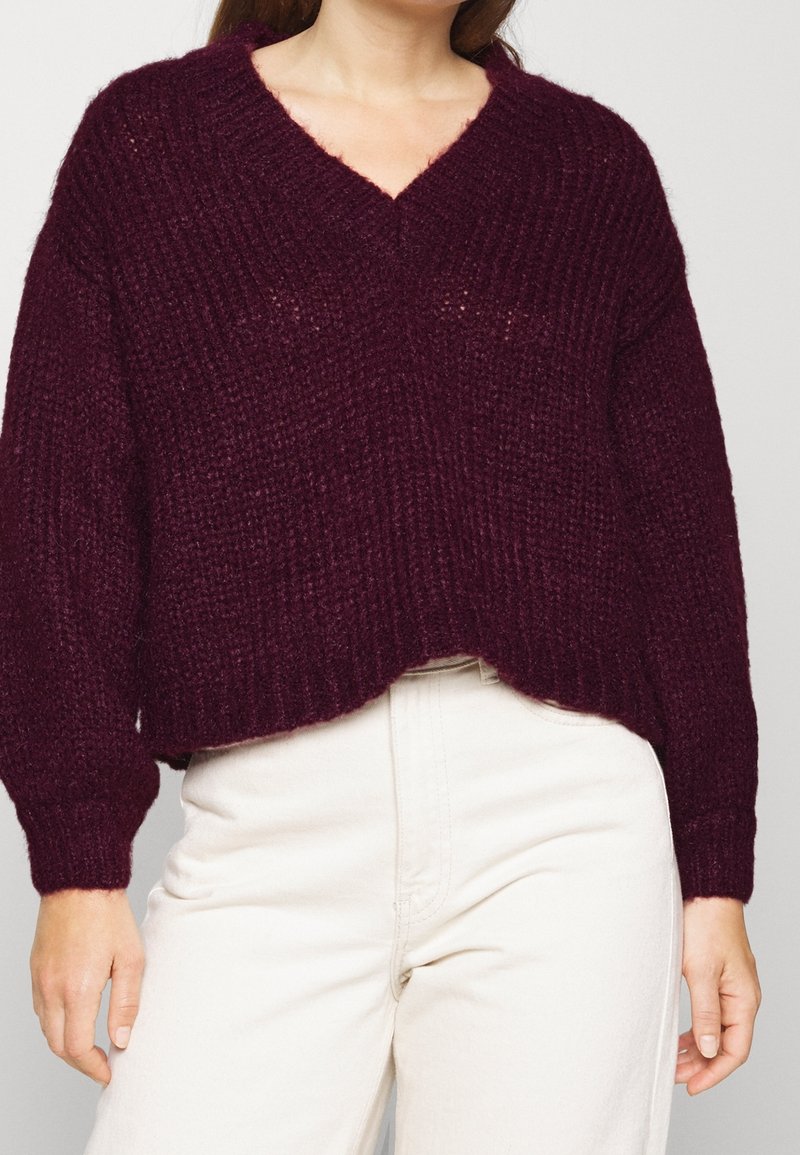 Burgundy stickad tröja med V-ringning, lös passform, ribbad struktur och långa ärmar, som matchas med ljusa byxor.