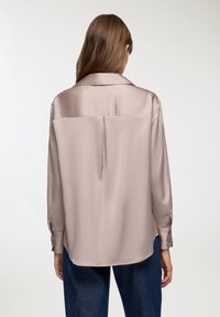 Camicia in satin color taupe chiaro con maniche lunghe, un pince sul retro e polsini con bottone. Presenta un colletto classico e una vestibilità rilassata.