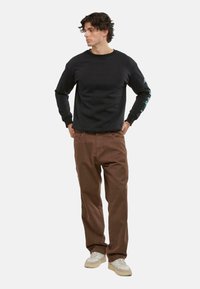 Felpa nera con colletto rotondo, maniche lunghe e dettaglio grafico turchese. Abbinata a pantaloni marroni e sneakers beige.