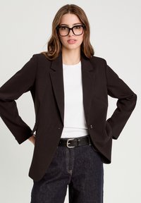 Blazer noir avec des épaules structurées, fermeture à un bouton, et un tissu lisse, porté sur un haut blanc et un pantalon en denim foncé.
