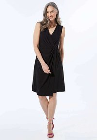 Schwarzes ärmelloses Kleid mit einem gedrehten Vorderdesign, knielanger Saum und aus glattem Stoff. Kombiniert mit fuchsia-farbenen High-Heel-Sandalen.