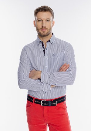 Mężczyzna z krótkimi włosami i brodą, ubrany w pasiastą koszulę na guziki oraz czerwone spodnie, stojący z założonymi ramionami na tle białego tła.