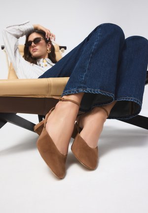 Mujer reclinada en una silla marrón claro, vistiendo vaqueros azul oscuro, zapatos marrones de ante con tacón bloque y correa al tobillo, camisa a rayas, gafas de sol y pendientes dorados.