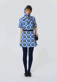 Žena u plavoj haljini s cvjetnim uzorkom i odgovarajućom maramom na glavi, žutim sunčanim naočalama, crnim tajicama i crnim cipelama, držeći crnu torbicu.