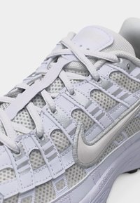 Κοντινή λήψη λευκού και ασημένιου αθλητικού παπουτσιού Nike με πλέγμα, κορδόνια και εμφανές ασημένιο λογότυπο swoosh στο πλάι.