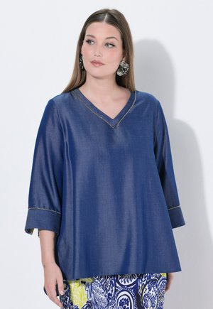 Donna con capelli castani lunghi che indossa una maglia blu a scollo a V e maniche a tre quarti, abbinata a pantaloni con motivo blu, bianco e giallo.