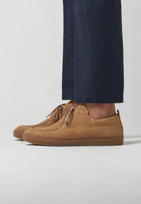Zapatillas de ante marrón con punta redonda, suela plana de goma y cordones. Los laterales presentan una marca minimalista y una textura suave.