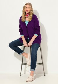 Lila kofta med svarta knappar, lagd över en blå topp. Mörk denimbyxor med uppvikta fållar och ljusrosa slitna skor.
