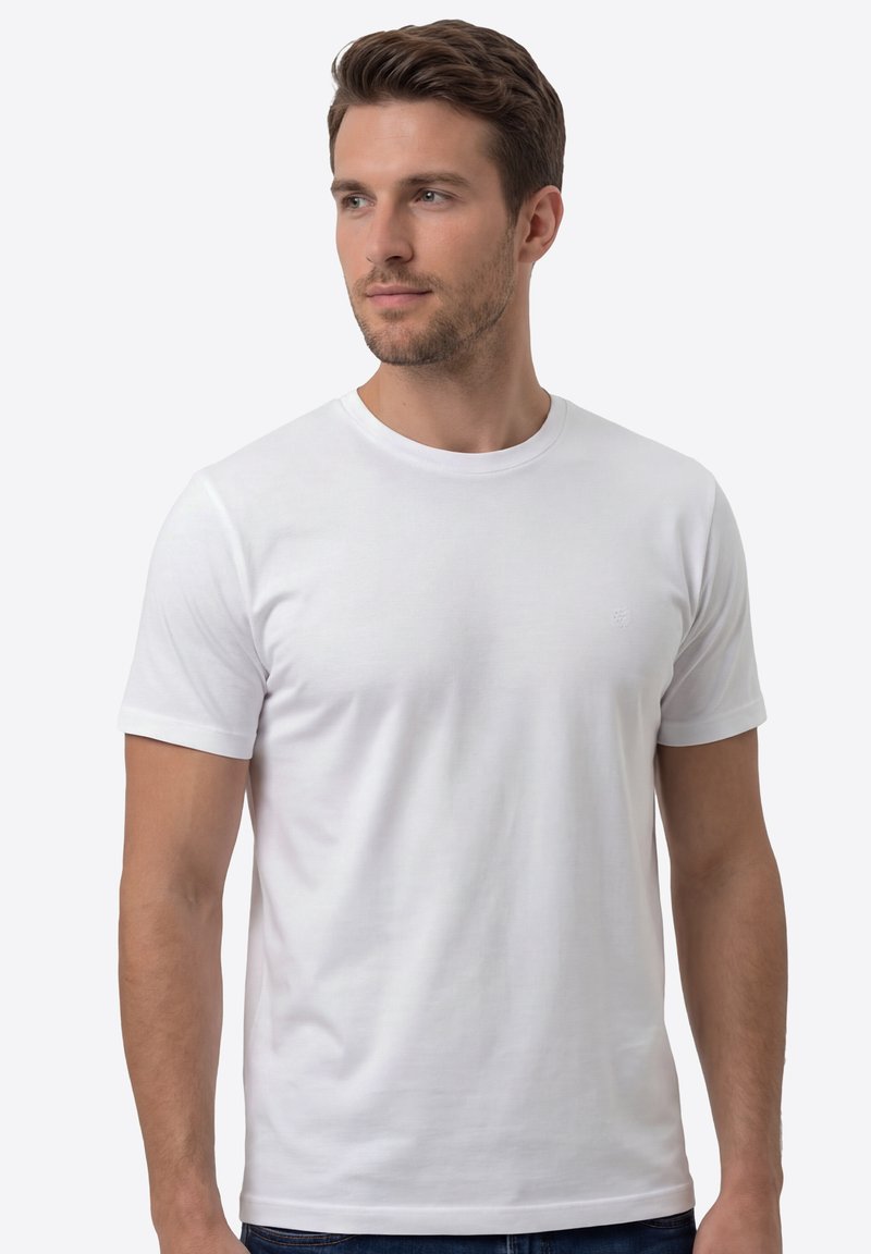 Homme aux cheveux courts et bruns portant un t-shirt blanc uni à manches courtes, regardant légèrement vers la gauche, sur un fond clair.