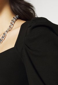 Svart ribbad topp med puffärmar och fyrkantig halsringning. Ett silverhalsband syns mot tygtexturen.