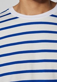 Gestreiftes Kurzarmhemd mit weißem Grund und kräftigen blauen horizontalen Streifen. Verfügt über einen runden Halsausschnitt und einen weichen, strukturierten Stoff.