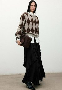 Mujer con suéter argyle marrón y beige, camisa blanca, falda negra larga con volantes, botas negras, sosteniendo un bolso marrón oscuro, de pie contra una pared blanca.
