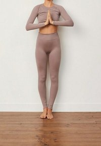 Jasnobrązowa dopasowana bluzka z długim rękawem oraz legginsy z wysokim stanem o prążkowanej fakturze, zestawione z bosymi stopami na drewnianej podłodze.