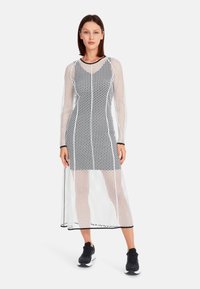 Durchsichtiges schwarz-weißes Kleid mit einem langen, lockeren Schnitt, das ein Netzdesign und lange Ärmel aufweist, kombiniert mit schwarzen Turnschuhen. Horizontale Saumlinie.