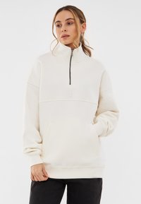 Cream-colored sweatshirt with a high collar and a front zipper, featuring a kangaroo pocket and ribbed cuffs. Smooth cotton fabric.