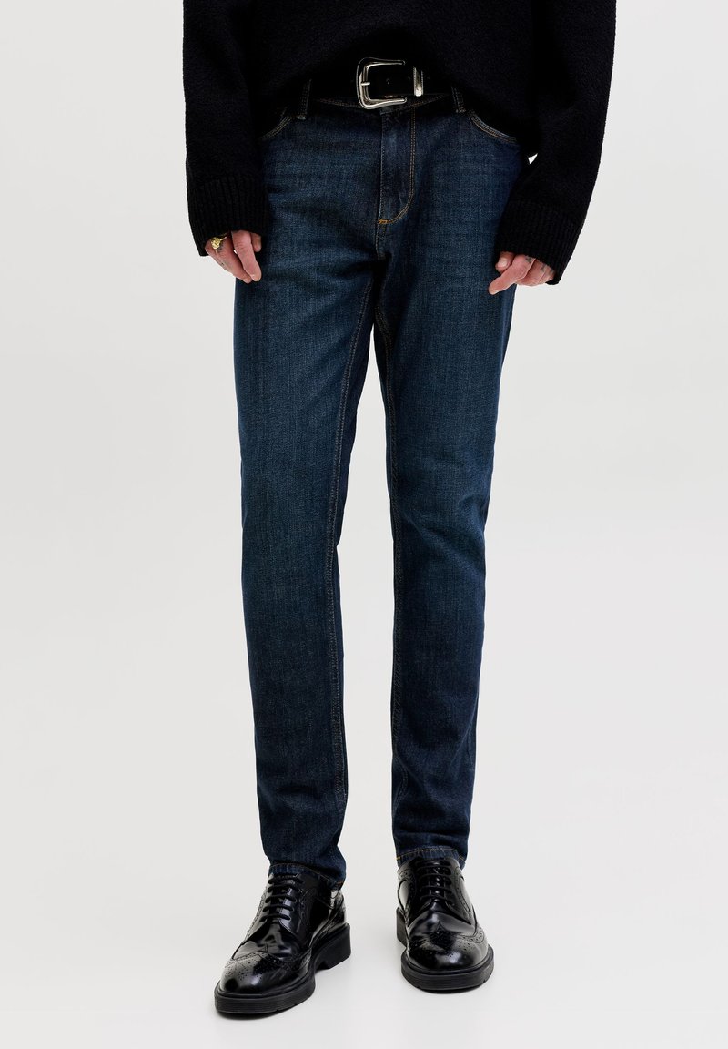 Personne portant un jean slim bleu foncé, une ceinture noire avec une boucle argentée, un pull en maille noire et des chaussures de ville noires brillantes.