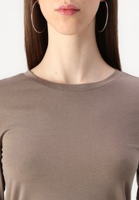 Barna, testhez Simuló felső, kerek nyakkivágással, puha anyagból készült. Finom textúrája van, enyhe fényével. Nagy ezüst karika fülbevaló látható.