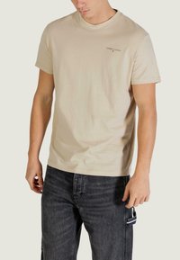 Beiges Baumwoll-T-Shirt mit Rundhalsausschnitt, kurzen Ärmeln und einem dezenten "TOMMY JEANS"-Logo auf der Brust. Kombiniert mit dunkelgrauen Jeans.