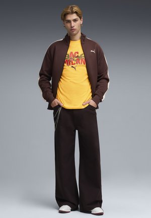 Młody mężczyzna w brązowej kurtce Puma, żółtej koszulce AC Milan, szerokich czarnych spodniach z łańcuchem oraz białych tenisówkach, stoi z rękami w kieszeniach.