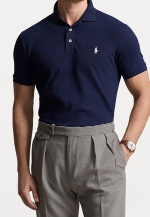 Man die een navyblauw poloshirt met wit logo, grijze, hooggetailleerde geplooide broek en een polshorloge aan de linkerhand draagt.