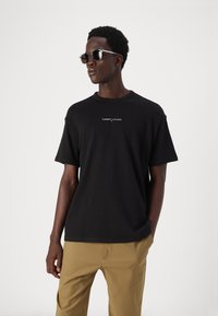 Černé bavlněné tričko s malým bílým logem na hrudi, doplněné khaki kalhotami a kulatými slunečními brýlemi. Jednoduchý design a střih.