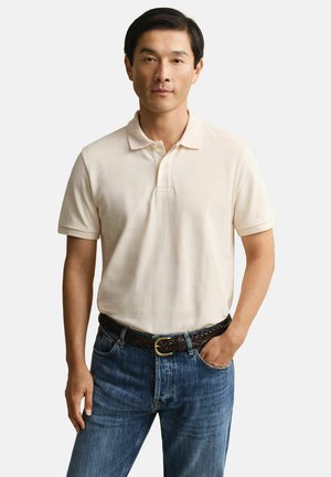 Homme portant un polo beige à manches courtes rentré dans un jean bleu, avec une ceinture tressée sombre, debout avec une main dans la poche.