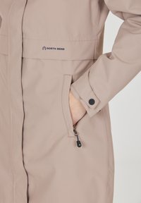 Lys beige vandtæt jakke lavet af tekstureret stof, med en forlomma, trykknap og snøre detaljer i kanten.