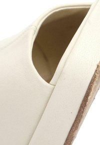 Gros plan d'une chaussure blanche à enfiler montrant la tige texturée, l'ouverture et une partie de la semelle avec un bord marron.