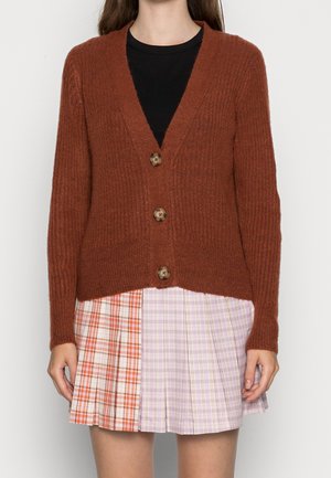 Vrouw draagt een roestkleurige, met knopen gesloten cardigan over een zwart topje en een geplooide minirok met oranje en roze geruite patronen.