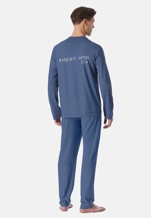Man staat op blote voeten, draagt een blauwe top met lange mouwen en een broek met de tekst "Morning Coffee Club" op de achterkant van de top.