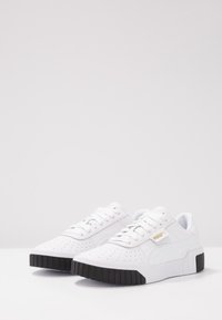 Baskets en cuir blanc avec un embout perforé, des lacets plats, un logo doré et une semelle en caoutchouc noire texturée.