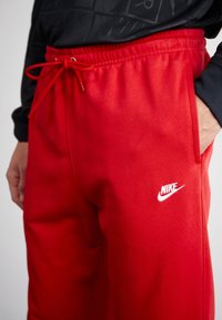 Röda Nike-byxor med elastisk midja, snörning, sidofickor och en vit logotyp på vänster lår. Mjuk, slät tygtextur.