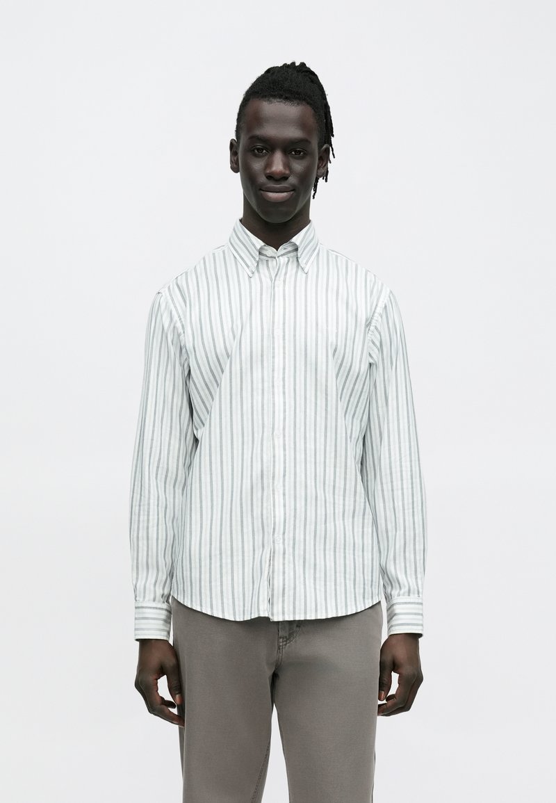 Raidallinen vaaleanvihreä ja valkoinen nappipaita, pitkähihaiset, kauluksellinen, valmistettu kevyestä kankaasta. Yhdistetty vaaleanharmaiden housujen kanssa.