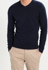 Pull à col en V bleu marine en tissu tricoté, avec manches longues et poignets et ourlet côtelés. Porté avec un pantalon beige clair.