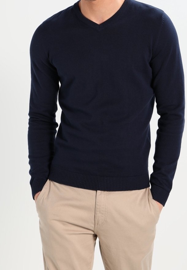 Pull à col en V bleu marine en tissu tricoté, avec manches longues et poignets et ourlet côtelés. Porté avec un pantalon beige clair.